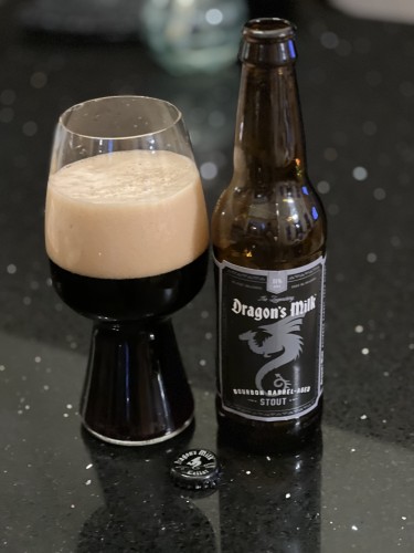 New Holland brewing Dragons Milk bourbon-barrel aged stout beer in a pint glass. The bottle label is black with silver and white lettering and a silver side view of a curled dragon with extended wings