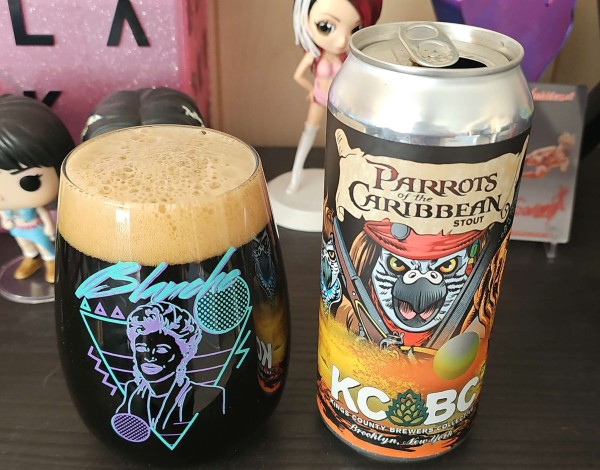 A glass filled with beer and a 16oz can sitting on a shelf. The glass features Blanche of the Golden Girls, with 80s like design behind her. The can features Parrots in Pirates of the Caribbean movie costumes 