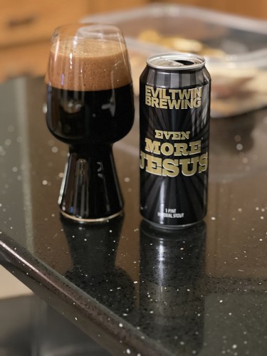 A stout glass filled with fudge dark beer and a deep brown head. A can of EvilTwin Brewing Even More Jesus into the left, black with bold gold lettering and a sunburst design.