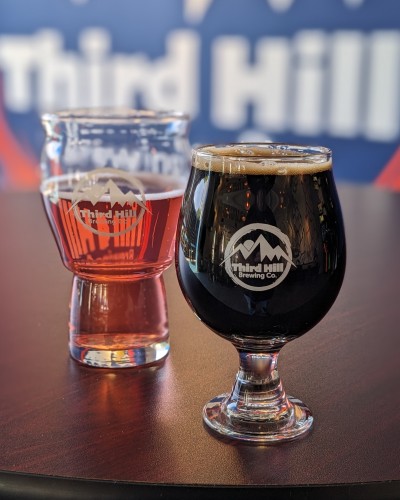 Left: Red clear Selzer in a glass.
Right: Black beer in a glass.
Both: Third Hill Brewing logo on glass.
Behind: Blurry Third Hill Logo on the wall.