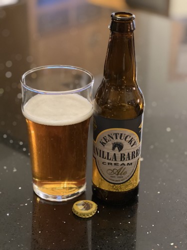 A pint glass of golden beer is to the left of its bottle, a yellow and white and black background with black and shiny gold lettering. The center of the label has a black horse head logo.