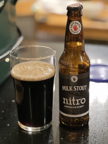 A 12 ounce bottle of LeftHand Brewing Milk Stout Nitro, with the iconic red left hand logo on the neck label and a black body label with white lettering. A pint glass of beer is to the left 
