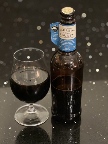Goose Island Bourbon County Biscotti stout in a tall brown bottle with a blue and tan label on its neck. A half full tulip glass of dark brown beer is to the left.