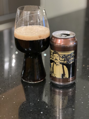 A stout glass of dark beer with a tan head is next to its can - dark brown with a tan and black stylized woodsman and the words “George Hunter Stout” in a scripted font