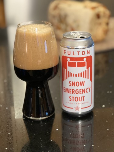 A can of Fulton Breeing Snow Emergency stout is on a white can with bright red lettering, in the style of a snow emergency plow route street sign. The image of a snow plow appears to be pushing the white background and lettering forward.

A stout glass with a significant tan head is to the left. A loaf of fresh baked bread is in the background.