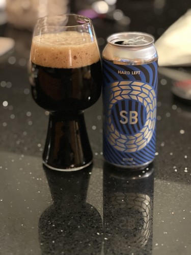 A silver can of Spiral Brewery Har Left Stout. The label is dark blue with black accents and silver lettering. A bold “SB” is in the middle of concentric silver circles.  A stout glass of dark beer with a tall tan head is to the left.