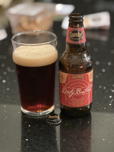 A bottle of founders Dirty Bastard scotch ale with a red label and tan lettering.  A pint glass of ruby red beer is to the left.
