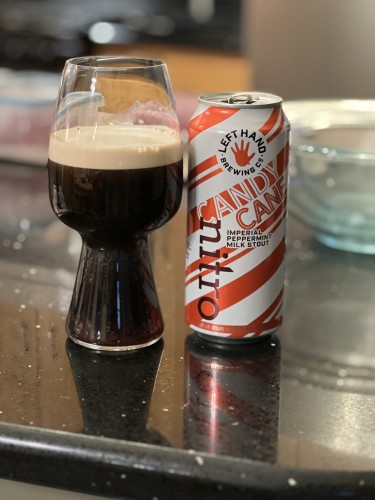 A candy cane striped pint can of beer, bright white and red diagonal stripes, and the iconic Left Hand Brewing red hand logo. A stout glass to the left holds the dark beer with a rich tan head.