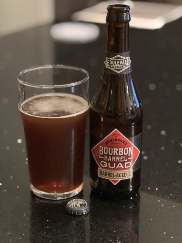 Boulevard Brewing Barrel-Aged Quad - a dark brown bottle with a red label with white and black lettering. A pint glass to the left holds the ruby-red beer