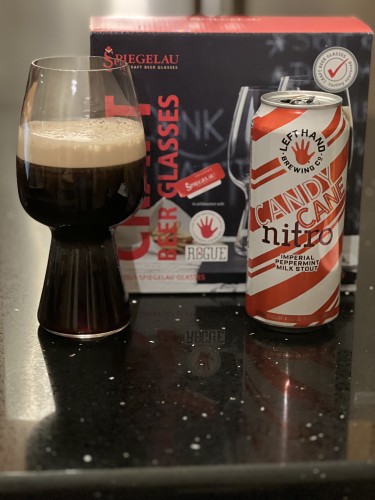 A can of Left hand Candy cane peppermint milk stout, shiny wrapped in angled white and red candy cane stripes. A special stout glass with a pinched middle is to the right. Behind is the box for the glasses showing the Left Hand logo