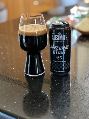 A black can with white writing holds AleSmith Speedway Stout beer. The can has a pair of crossed checkered finish line flags in the center. A stout glass full of beer with a tan head is to the left.
