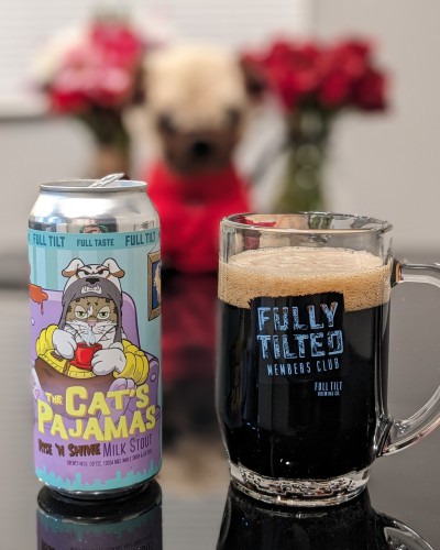 Left: Can of beer. Label features a drawing of a cat in pajamas on a couch.

Right: A glass mug that says Fully Tilted Members Club. Black beer inside.

Behind: Out of focus flowers and a stuffed dog.