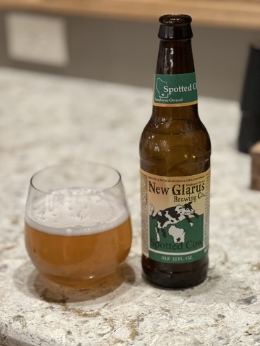 A brown bottle of New Glarus spotted cow beer, with a green neck label and a yellow body label with green grass and a black and white spotted cow leaping over the state of Wisconsin. A wine glass with the light golden ale is to the left