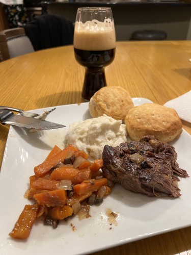 A plate with orange carrots, white mashed potatoes, two golden brown biscuits, and brown roasted meat. In the background is a stout glass of Central Waters Satin Solitude beer with a rich tan head