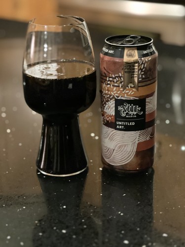 A stout glass of deep dark beer with the can from Untitled Art to the right. The can is artfully decorated in shades of brown and white and gold with a shiny gold pendant reading “barrel aged”