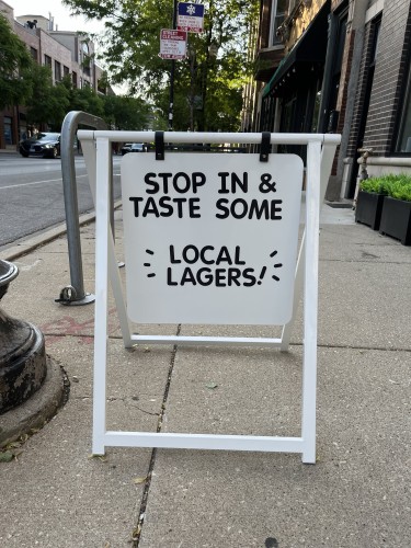A sidewalk sign outside a building reads, "STOP IN & TASTE SOME LOCAL LAGERS!" In the background, there is a street with buildings, trees, and a parking sign.