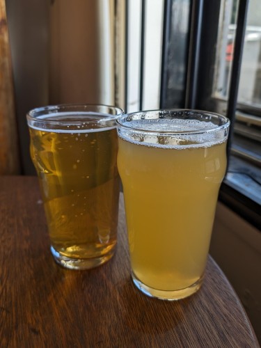 Two pints of beer on a dark wooden surface in front of a window. The beer on the left is clear while the one on the right is a hazy pale.